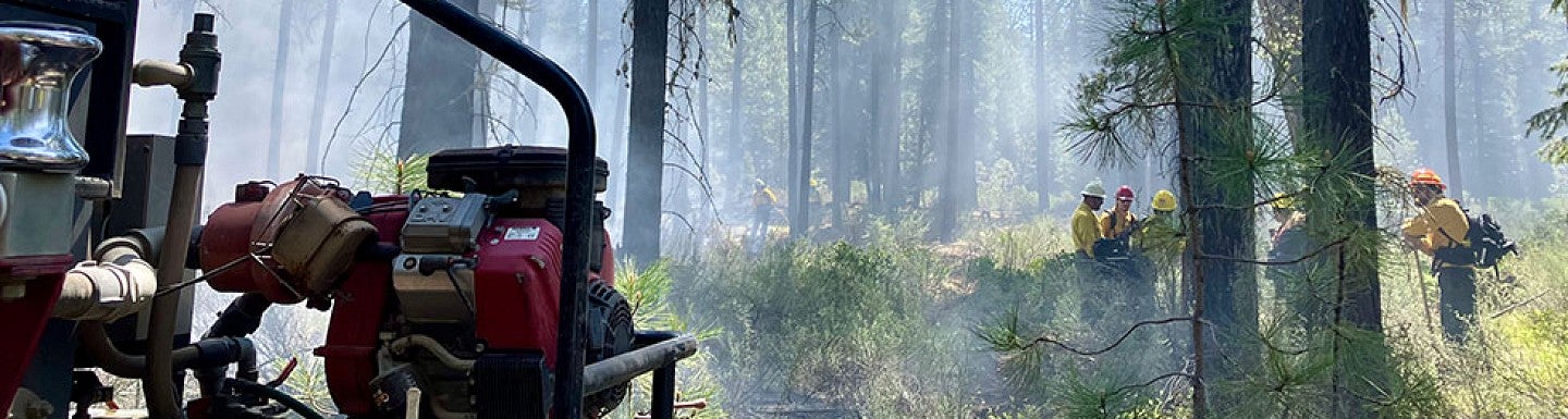 Firefighters in yellow protective gear work amidst smoke in a forest, with sunlight filtering through tall trees. In the foreground, a red water pump and hoses are visible, indicating active fire control efforts.