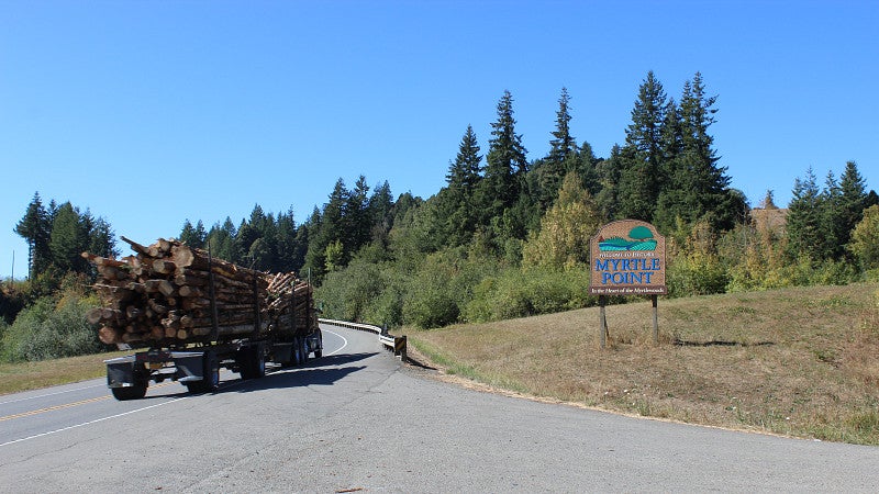 Logging truck driving down the road past a welcome sign for Myrtle Point, Oregon.