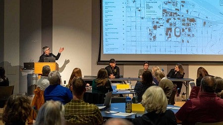 Person giving a presentation at a podium pointing to large map with seated panel to one side in front of 